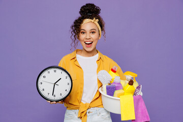 Young surprised shocked happy housekeeper woman wear yellow shirt hold basin with detergent bottles clock tidy up show time isolated on plain pastel light purple background studio. Housework concept.