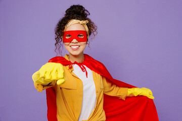 Young housekeeper woman wear yellow shirt rubber gloves red cloak mask do super hero gesture stretch hand to camera tidy up isolated on plain pastel light purple background studio. Housework concept.