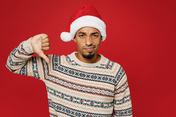 Merry sad young man wear warm cozy Christmas sweater Santa hat posing showing thumb down dislike gesture isolated on plain red color background studio. Happy New Year 2023 celebration holiday concept.