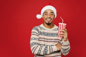 Merry young man wear warm cozy Christmas sweater Santa hat posing look camera hold cup of soda pop...