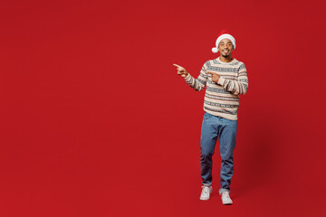 Fototapeta premium Full body merry fun young man wear sweater Santa hat posing point index finger aside indicate on workspace area copy space mock up isolated on plain red background Happy New Year 2023 holiday concept
