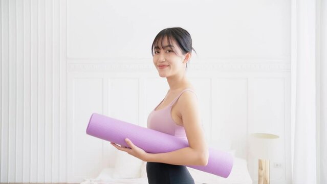 Asian woman turns and smiles at the camera, wearing sports bra and yoga pants holding a rolled-up yoga mat standing in a bedroom with sunlight from the window. Real time portrait video.