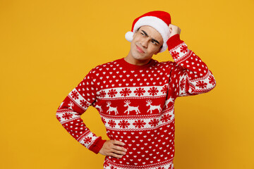 Sad young man wear red knitted christmas sweater Santa hat posing hold scratch head look aside on workspace area isolated on plain yellow background. Happy New Year 2023 celebration holiday concept.