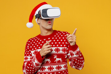 Merry young man wear red knitted christmas sweater Santa hat posing watching in vr headset pc gadget point finger aside on area isolated on plain yellow background Happy New Year 2023 holiday concept.