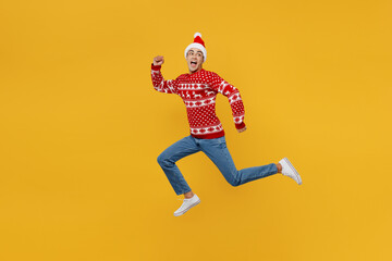 Full body side view merry young man wear red knitted Christmas sweater Santa hat posing run look back aside on area isolated on plain yellow background Happy New Year 2023 celebration holiday concept
