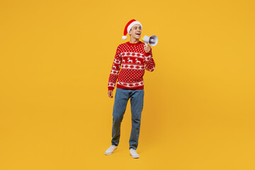 Full body merry young man in red Christmas sweater Santa hat posing hold scream in megaphone announces discounts sale Hurry up isolated on plain yellow background Happy New Year 2023 holiday concept
