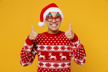 Merry satisfied fun young man wear knitted christmas sweater Santa hat glasses posing show thumb up...