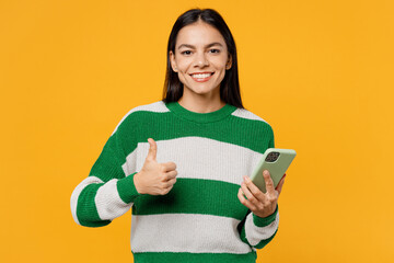 Young fun latin woman wear casual cozy green knitted sweater hold in hand use mobile cell phone in mint case show thumb up isolated on plain yellow background studio portrait People lifestyle concept.