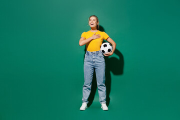 Full body young woman fan in basic yellow t-shirt cheer up support football sport team hold soccer ball watch tv live stream put hand in heart sing hymn song isolated on dark green background studio - obrazy, fototapety, plakaty