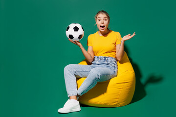 Naklejka na ściany i meble Full body young woman fan wear basic yellow t-shirt cheer up support football sport team sit in bag chair hold soccer ball watch tv live stream spread hands isolated on dark green background studio.