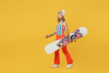Side view snowboarder fun woman wear orange suit goggles mask hat ski costume swimsuit spend extreme weekend walk go isolated on plain yellow background studio. Winter sport hobby trip relax concept.