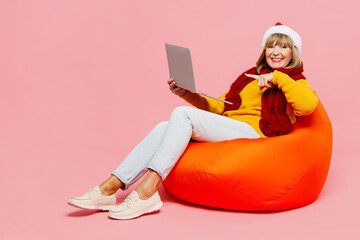 Full body elderly woman 50s year old wear yellow sweater red scarf Santa hat posing sit in bag chair work use point on laptop pc computer isolated on plain pink background Happy New Year 2023 concept