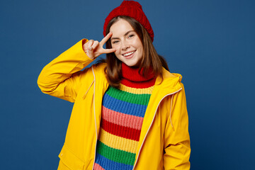 Young fun woman wear sweater red hat yellow waterproof raincoat outerwear showing cover eye with victory sign isolated on plain dark royal navy blue background Outdoor wet fall weather season concept