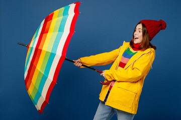 Side view young woman wear sweater red hat yellow waterproof raincoat outerwear try to hold opened umbrella isolated on plain dark royal blue background Outdoors wet windy fall weather season concept. - obrazy, fototapety, plakaty