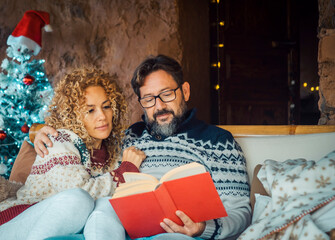 Serene young mature couple at home enjoying relax and indoor leisure activity in december holiday. Christmas tree in background. Man reading a book with a woman. Married together relationship love