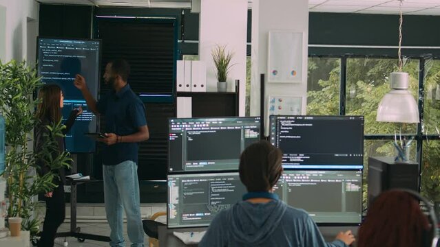 Diverse team of software engineers analyzing code on wall screen tv, using digital tablet and working with artficial intelligence. Coders parsing cloud computing project with big data.