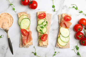 Tasty crispbreads with prosciutto, cream cheese and vegetables on white marble table, flat lay