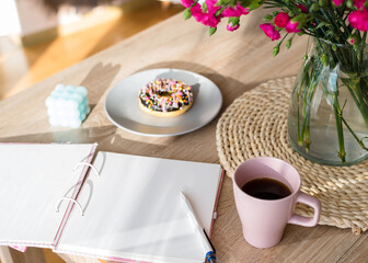 Cozy breakfast and planning. Notepad and pen, cup of coffee, pink donut, flowers and candles on the table. Women's desktop. Top view