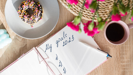 My goals 2023. Handwritten text in a notebook on the table and a pen, a cup of coffee, a pink donut, flowers and candles on the table. Girl's plans for 2023.