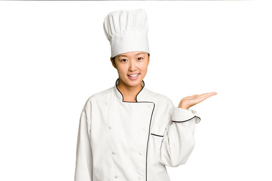 Young asian cook woman isolated showing a copy space on a palm and holding another hand on waist.