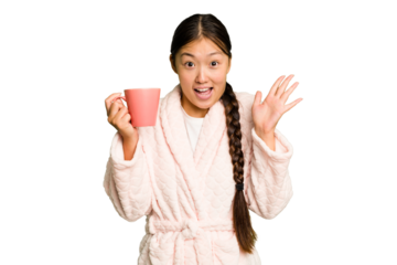 Young asian woman wearing a pajama holding a cup isolated receiving a pleasant surprise, excited and raising hands.