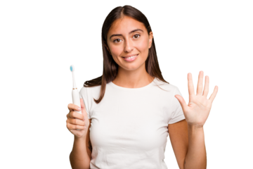 Young caucasian woman holding an electric toothbrush isolated smiling cheerful showing number five with fingers.