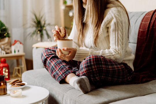 Cozy woman legs in knitted winter warm socks, sweater and checkered plaid drinking hot cocoa or coffee in mug, during resting on couch at home. Christmas holidays with candle, decor and cookies