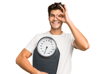Young caucasian man holding a scale isolated excited keeping ok gesture on eye.