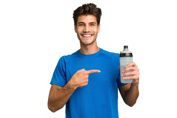 Young sport caucasian man holding a bottle of water isolated smiling and pointing aside, showing...