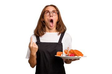 Young caucasian woman holding a waffles dish isolated pointing upside with opened mouth.