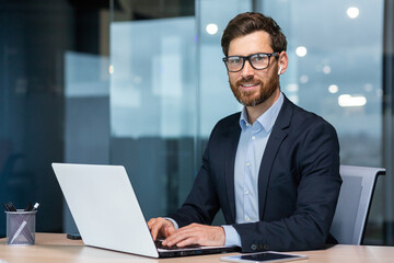 Portrait of mature successful and happy businessman, senior man with beard smiling and looking at camera, investor working with laptop inside office in business suit.