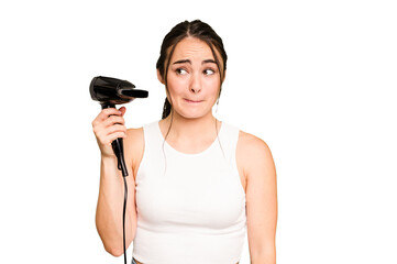 Young caucasian woman holding a hairdryer on green chroma background confused, feels doubtful and unsure.