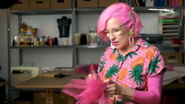 Tailor with pink hair and colorfull clothes hand sewing a piece of fabric in a sewing workshop.