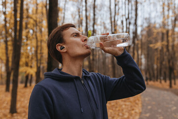 Sportsman drinking water from plastic reusable bottle while having break during training in autumn park 