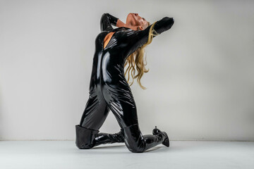 Blonde Fetish Figure Model Poses In A Studio Environment Wearing A Shingy One Piece Black Leather...