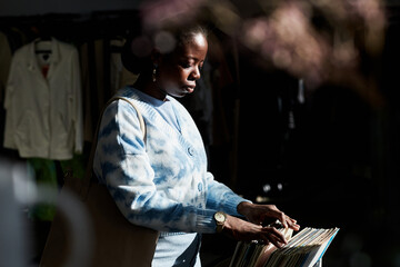 Moody side view portrait of black young woman choosing vintage records in thrift store lit by sunlight, copy space
