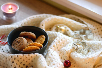 Cups of tea or coffee, bowl of cookies, soft blanket, lit candles, books and various Christmas decorations on the table. Selective focus.