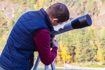 Man sets up telescope on tripod to survey mountains, autumn landscape and stars in night sky