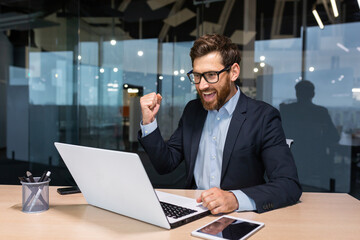 Successful businessman celebrating victory and good business achievement, boss using laptop reading notification from screen and holding hand up smiling triumph gesture.