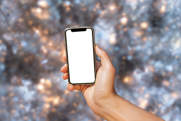 Mobile phone with empty white screen in hand on Christmas bokeh background. New phone gift for Christmas on a blurred white tree background