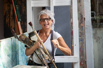 older gray haired mature happy artist woman with glasses and big paintbrushes make fist power...