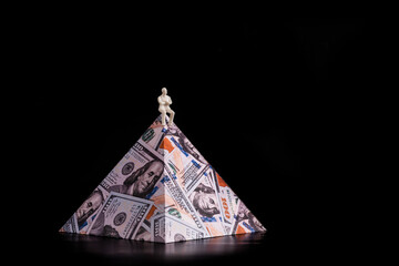 The symbolic figure of a businessman sits on top of a pyramid depicting 100 US dollar bills.