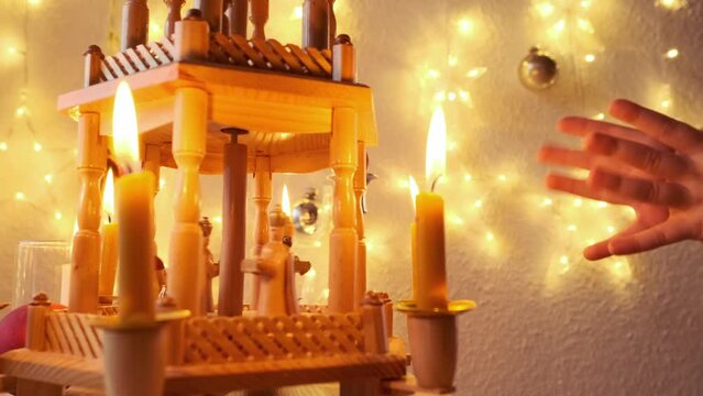 Christmas pyramid rotates with nativity scene and wood carving Christmas characters, traditional german christmas decoration, burning candle in dark room, children's palms clap joyfully