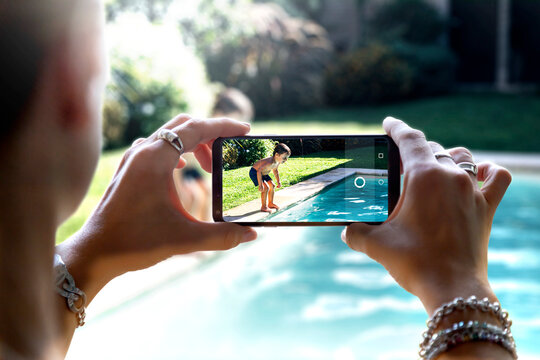 Woman taking a photo with a smartphone of a little boy jumping into a swimming pool.