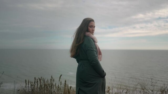 Back view of mixed ethnic woman standing on cliff, looking to sea view, turns to camera on moody cloudy day. Thoughtful long haired female in green coat overthinking near ocean. Wind blows hair.