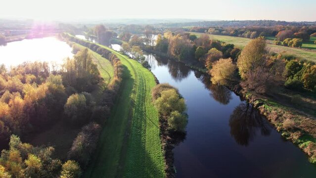 Aerial footage of a Still flowing river with mirror like sky reflection, with grass pathway, walkway and a trees.