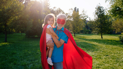 In a sunny day in the middle of the park posing in front of the camera grandfather and his niece in a superhero suits