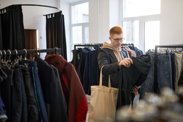 Wide angle view of young man browsing clothes on racks while shopping sustainably in thrift shop
