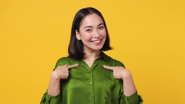 Beautiful friendly charming confident young woman of Asian ethnicity 20s she wear green shirt pointing fingers on herself blink wink eye isolated on plain yellow color wall background studio portrait