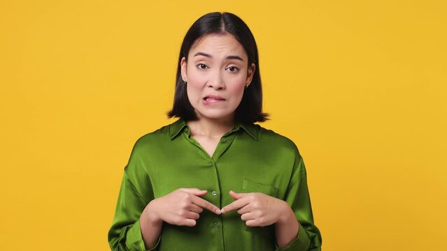 Fun confused shy portrait young woman of Asian ethnicity 20s she wear satin green shirt feels doubtful spreading hands say oops ouch oh omg i am so sorry isolated on plain yellow color wall background